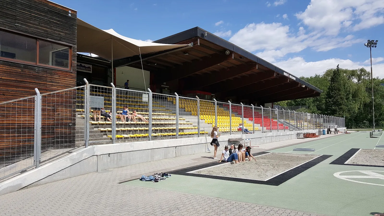 The side of the Brixen ice hall with its covered stand, wooden cladding and the outdoor paving in front. — Eishalle Brixen (Stadio del ghiaccio Bressanone)