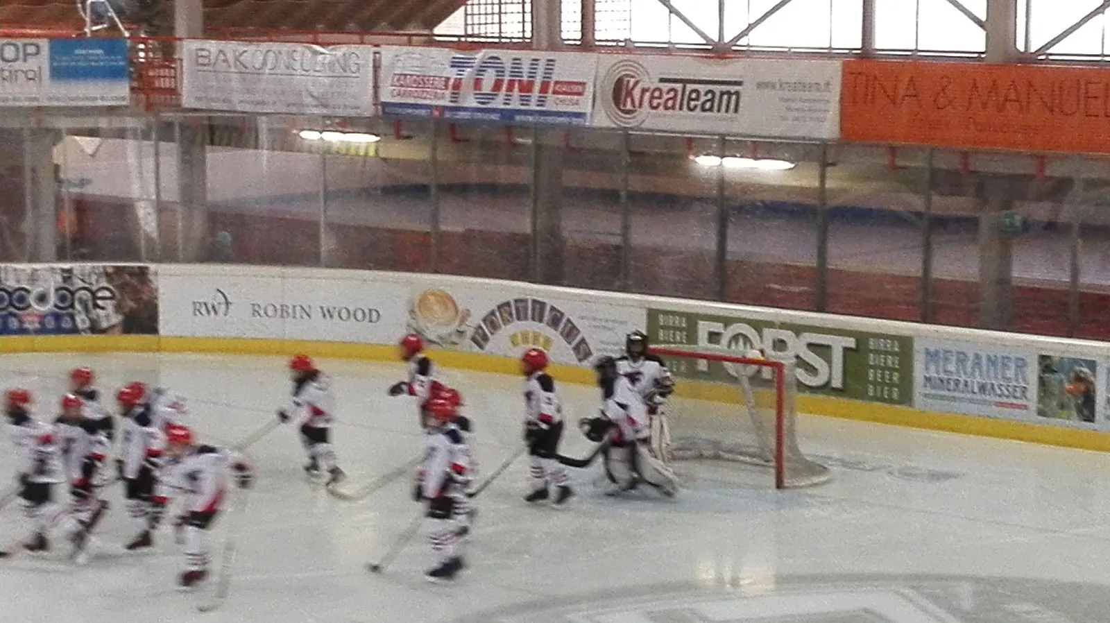 Young hockey players skating in a line along the boards of the ice hall, with sponsor panels on the dasher and the roof structure above. — Eishalle Brixen (Stadio del ghiaccio Bressanone)