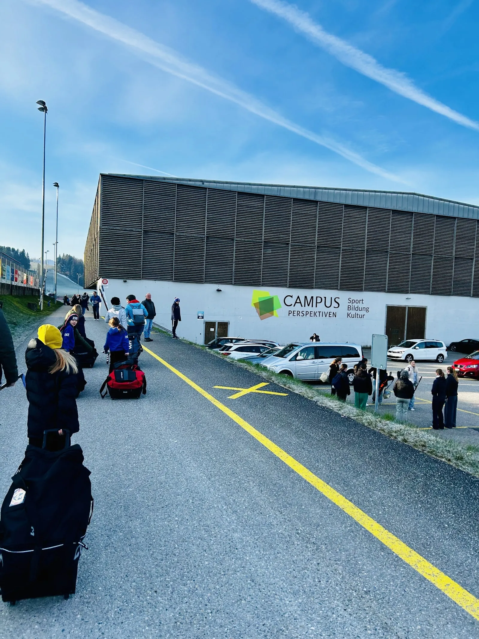 The side of the Campus Perspektiven building in Huttwil, with its slatted wooden upper wall, the Campus logo and parked cars, as seen from the access road. — Eishalle Campus Perspektiven