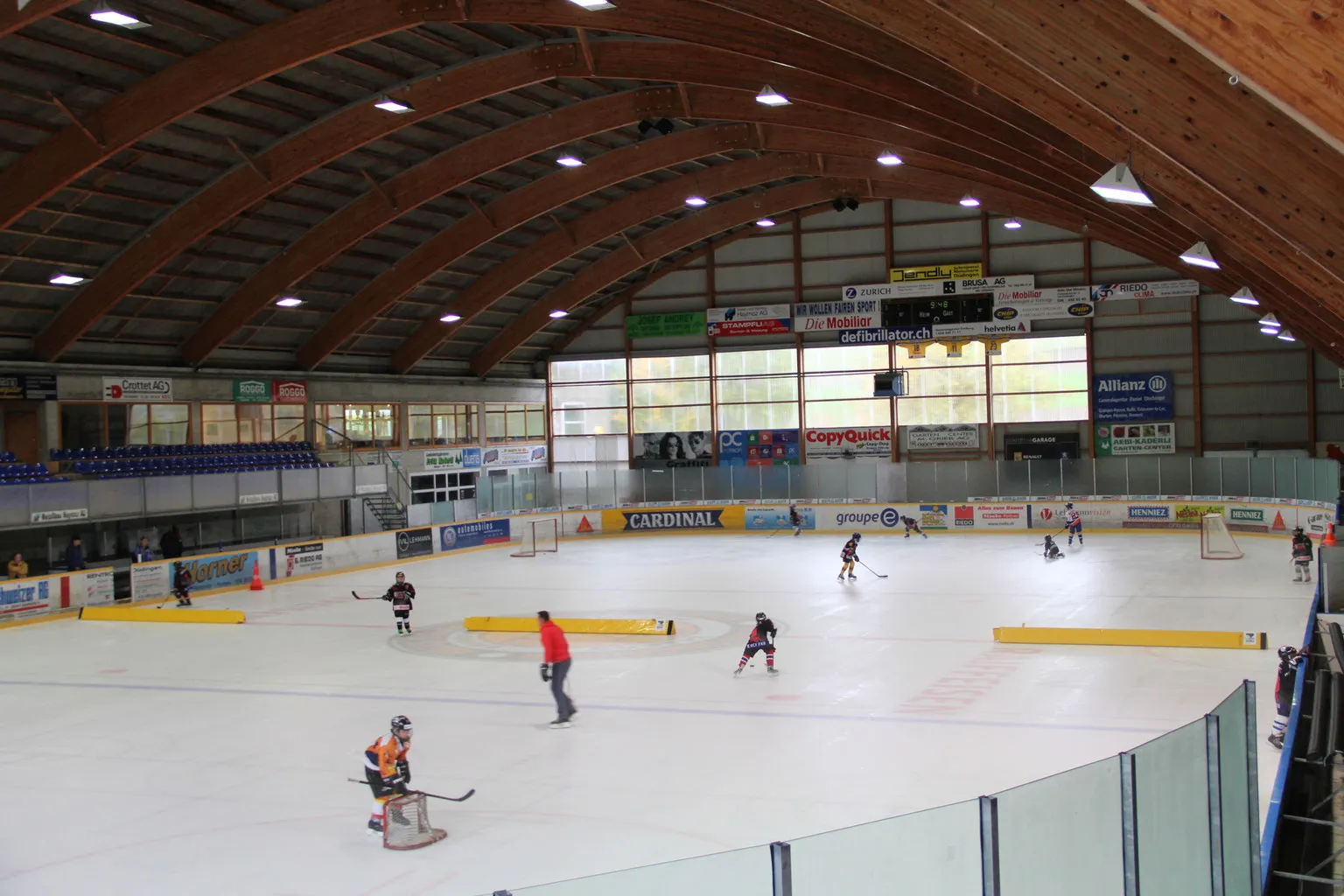 Die Eisfläche der Eishalle Düdingen unter ihrem geschwungenen Holzdach, mit einigen trainierenden Spielern am anderen Ende – Eishalle Düdingen