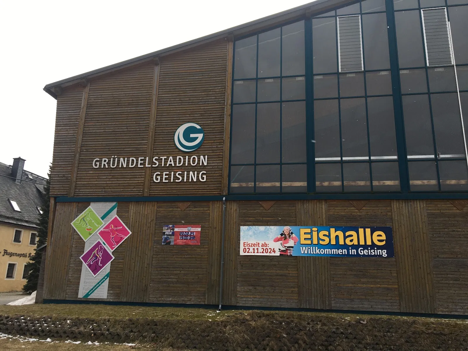 The timber-clad end wall of the hall with the Gründelstadion Geising lettering, a tall glazed gable and an Eishalle welcome banner. — Eishalle Gründelstadion Geising
