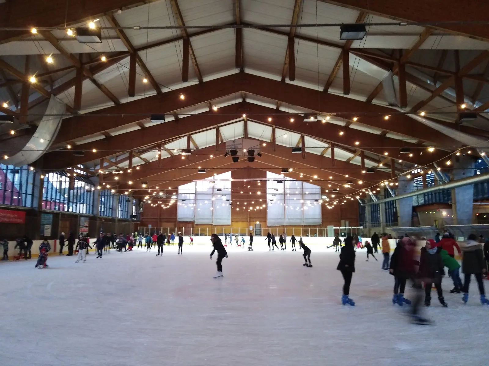A public session on the ice beneath the hall's timber roof trusses hung with strings of lights, with large gable windows at the far end. — Eishalle Gründelstadion Geising