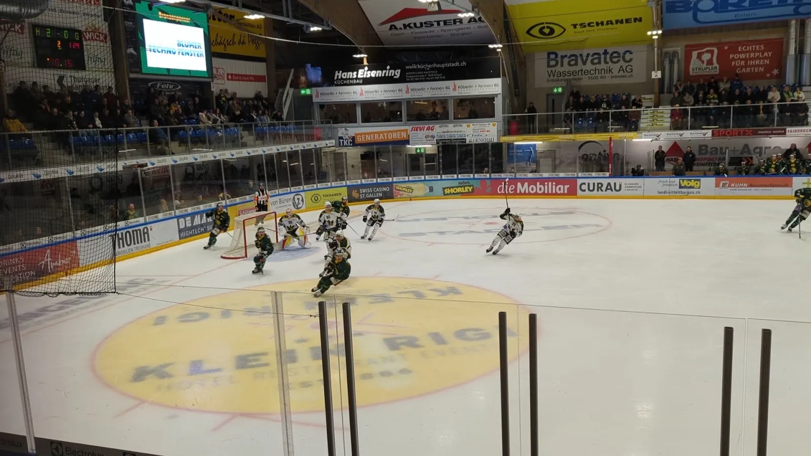 A hockey game in play in the Güttingersreuti hall in Weinfelden, players spread across the ice under the curved timber roof with the stands behind the netting. — Eishalle Güttingersreuti