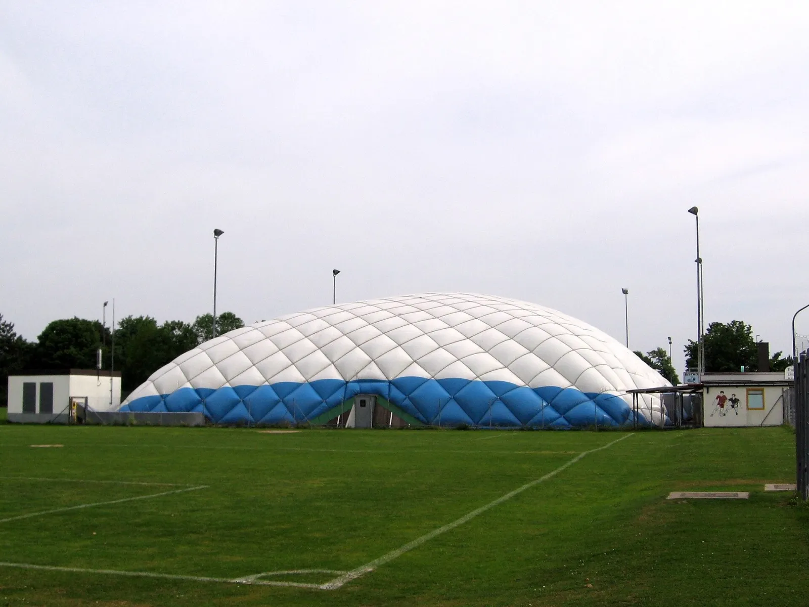 The white and blue inflated roof of the Haunstetten ice hall seen across a grass pitch, with floodlight masts around it. — Eishalle Haunstetten