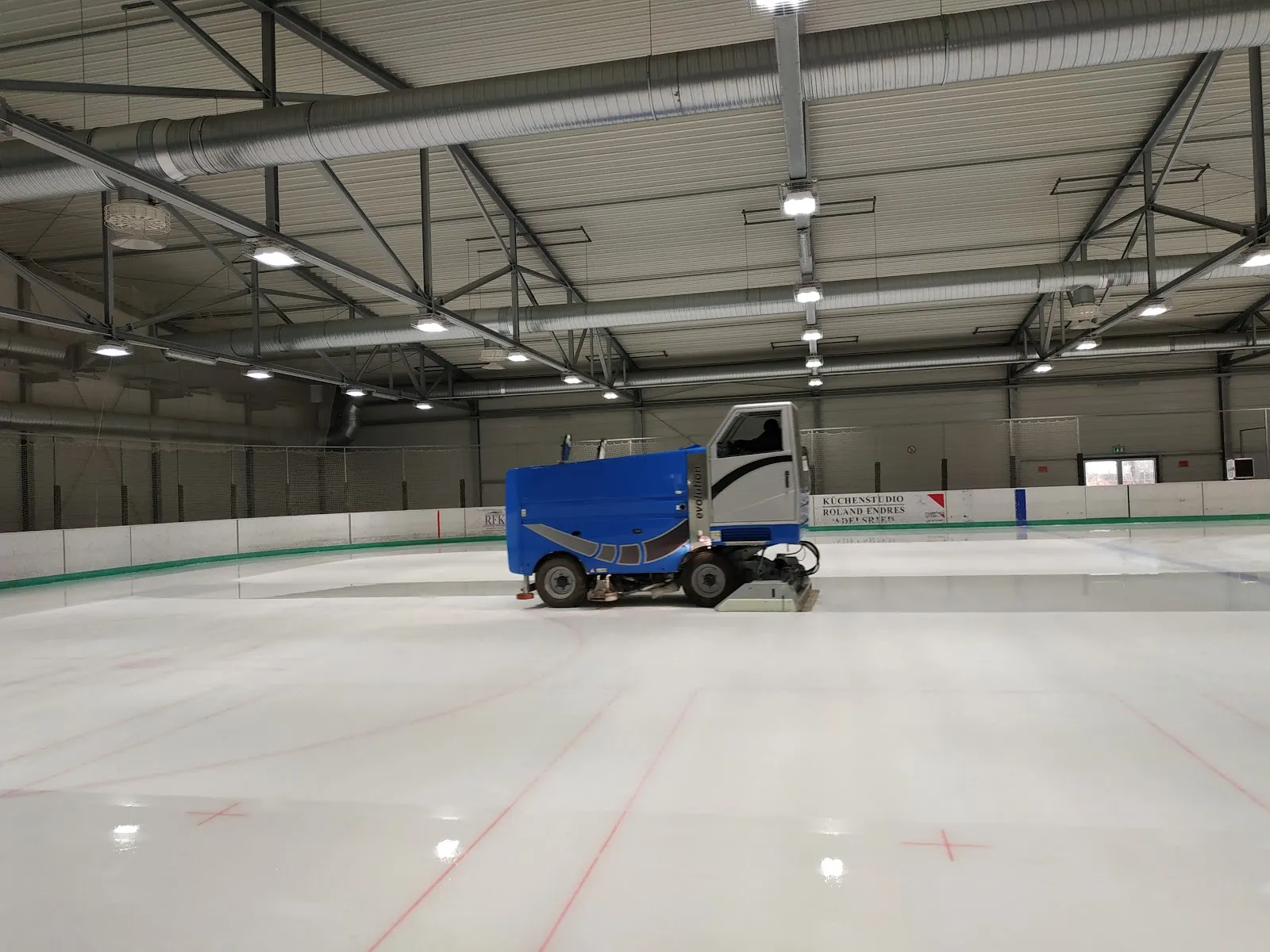 The empty, freshly resurfaced ice with red line markings, a blue ice resurfacer parked by the far boards and steel roof trusses above. — Eishalle Haunstetten