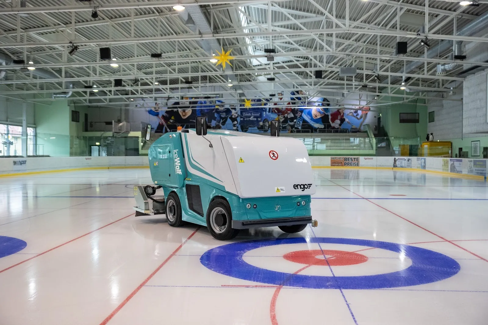 The ice surface of the Ilmenau Eishalle with a resurfacer crossing a curling target, the steel roof trusses and the board advertising behind. — Eishalle Ilmenau