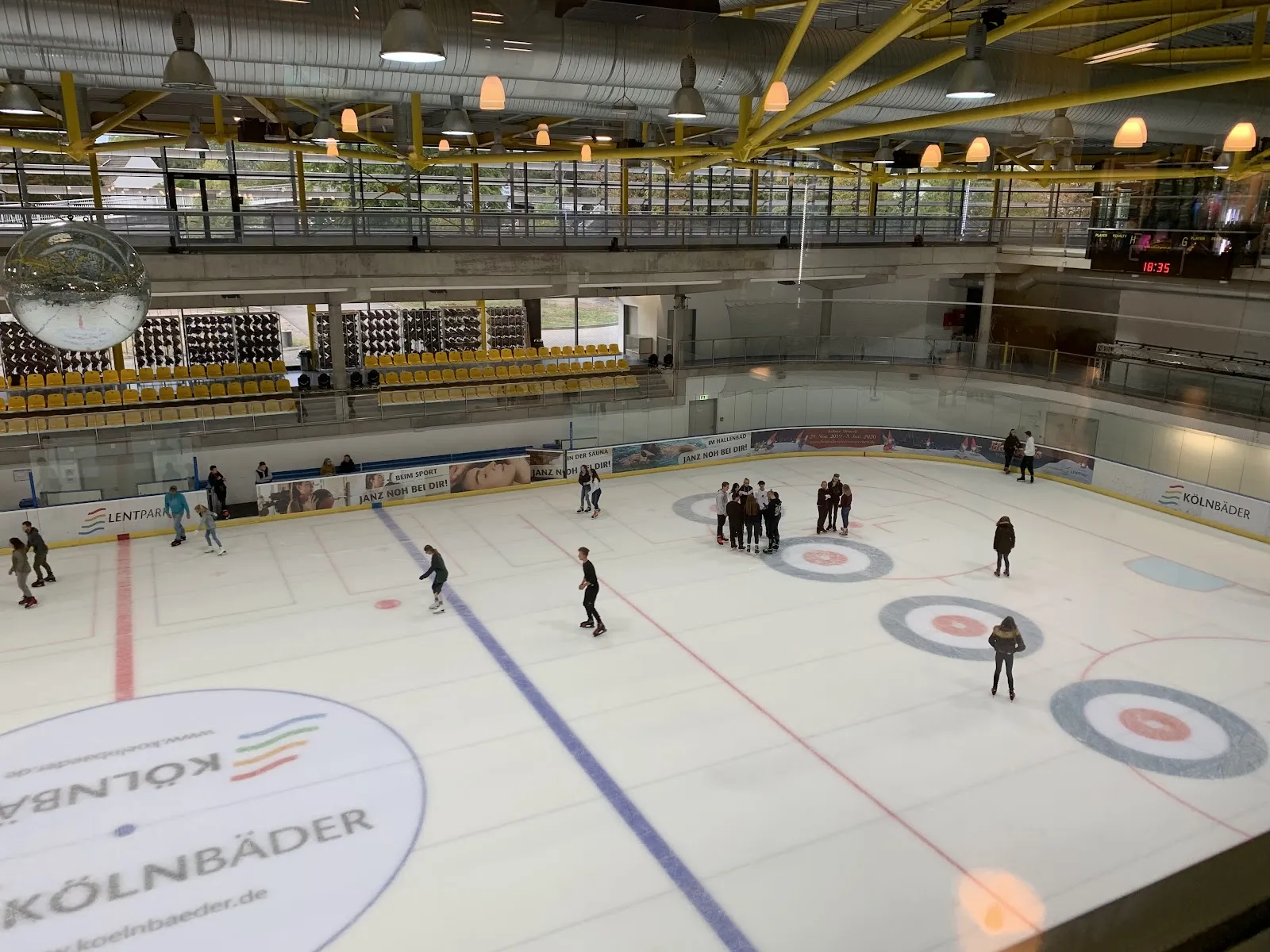 Läufer verteilt über die ganze Eisfläche der Lentpark-Halle in Köln, darüber die gelben Stahlbinder des Dachs und die Galerie der Eishochbahn. – Eishalle Lentpark