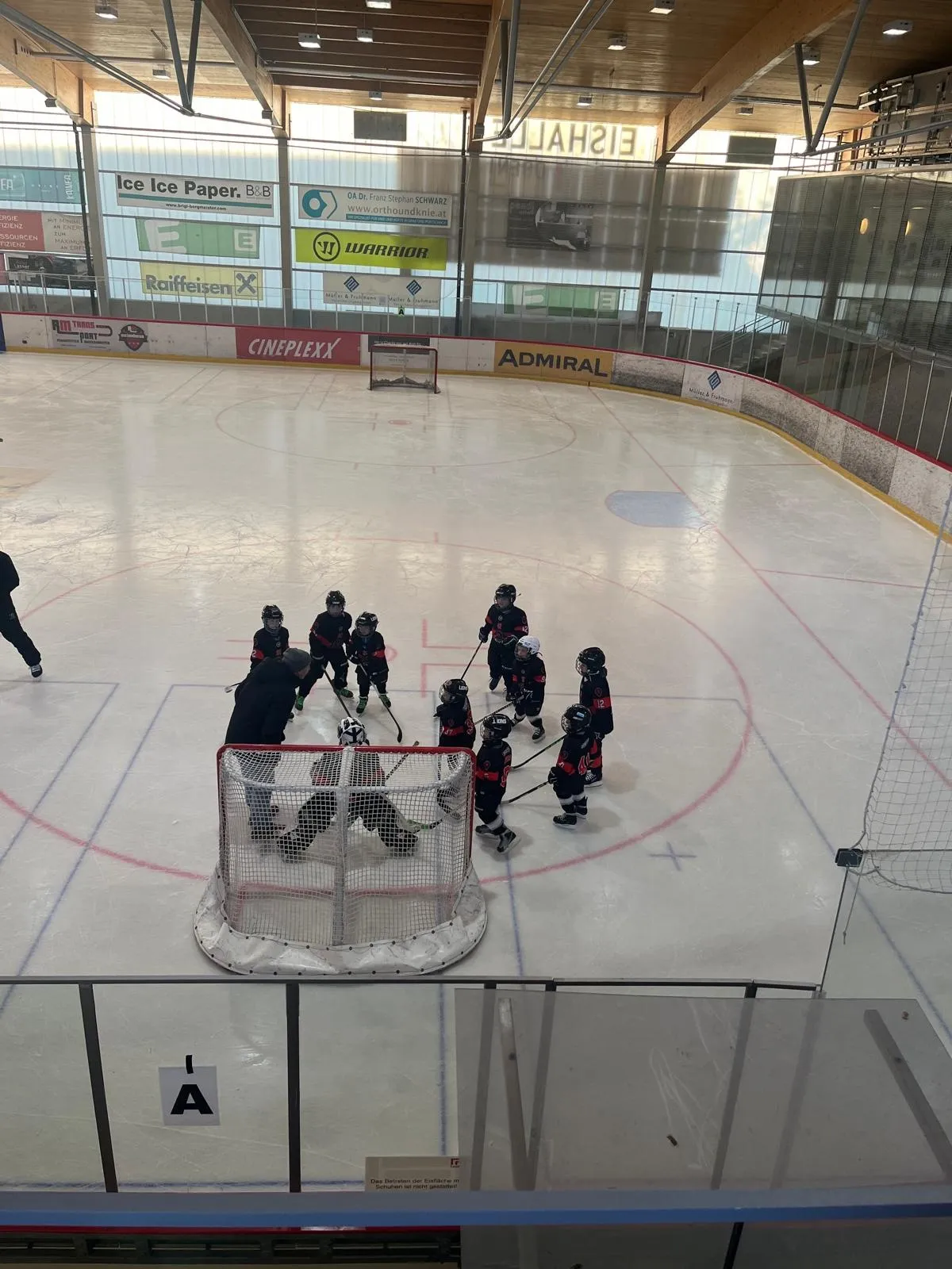 The ice at the Eishalle Leoben seen from the rink-side rail, with children practising round a goal and the timber roof, glazed upper wall and sponsor boards behind. — Eishalle Leoben