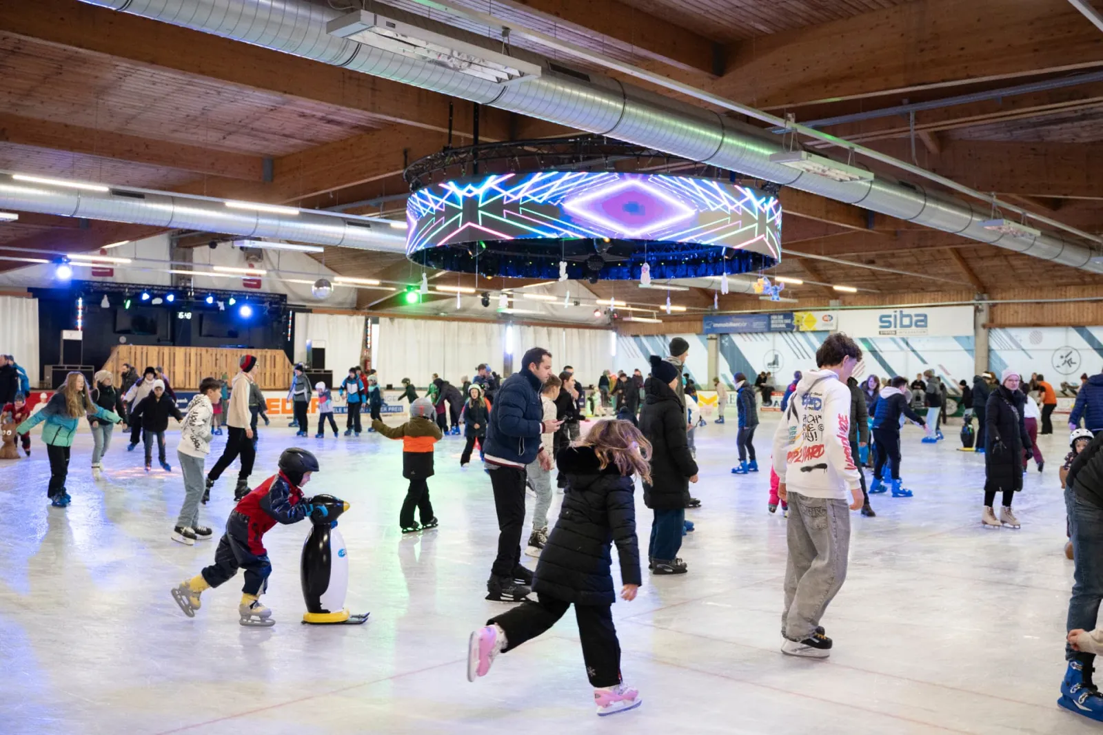 A busy public skating session in the timber-roofed hall: children and adults spread across the ice, with a ring of lights hanging over the rink. — Eishalle Peuerbach