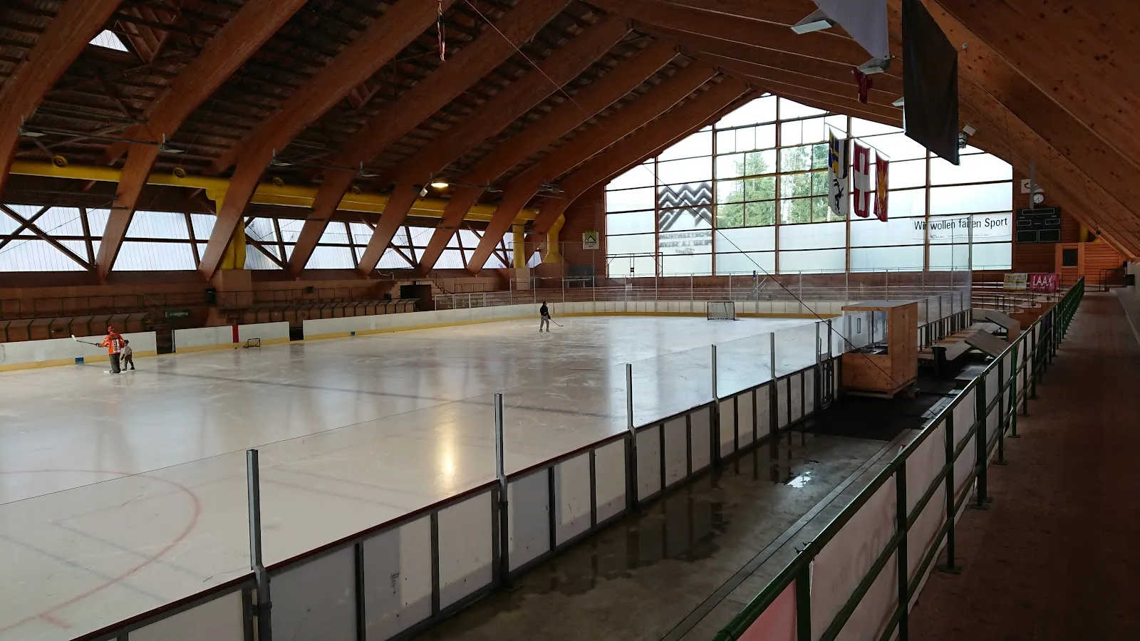 The covered ice hall seen along the boards from the stands, an empty ice surface under a curved timber roof with a few skaters at the far end. — Eishalle Sportzentrum Prau la Selva