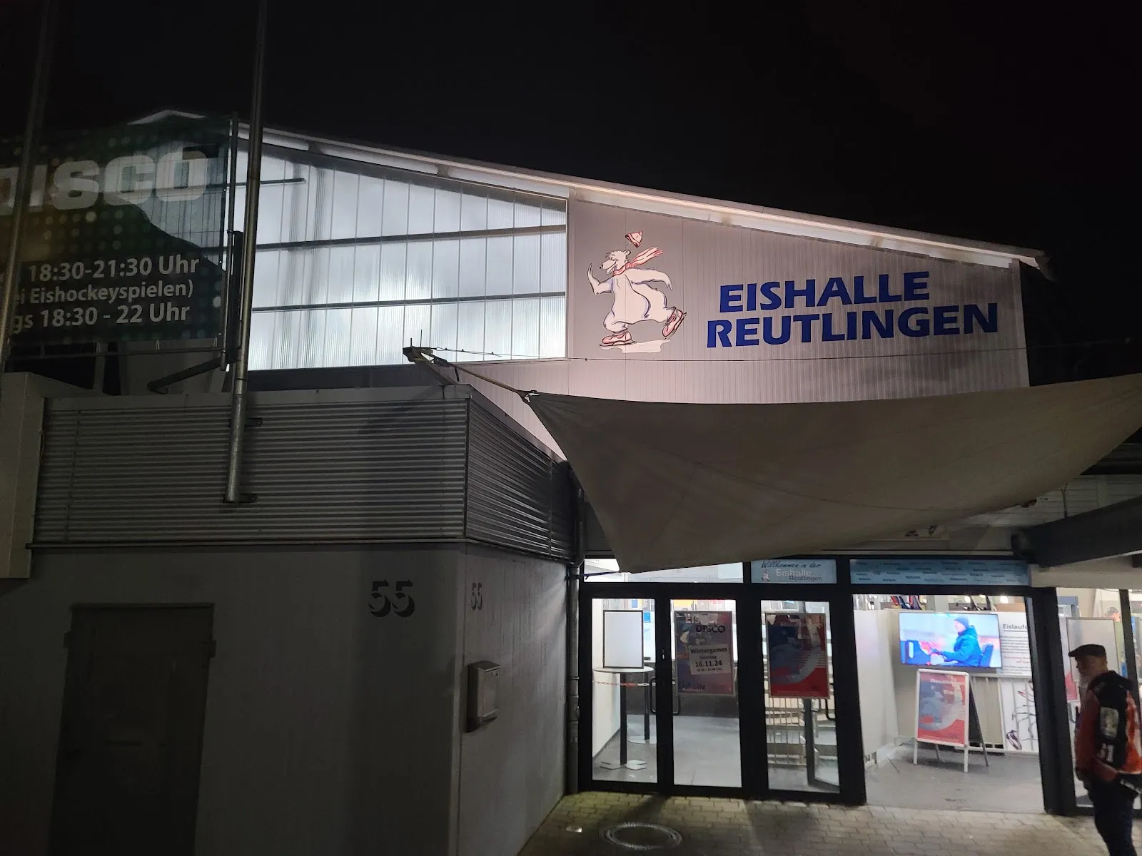 The entrance of the Eishalle Reutlingen at night, with the hall's name and ice-bear logo on the lit wall above the glass doors — Eishalle Reutlingen