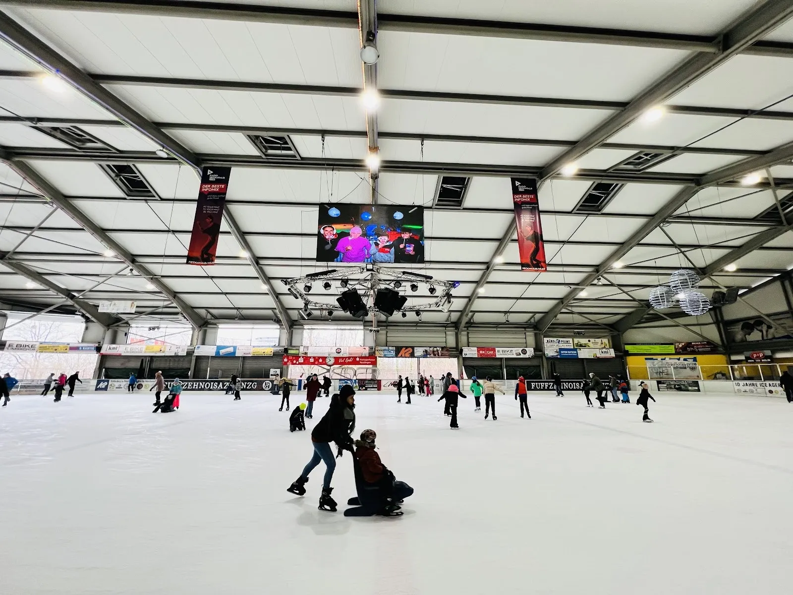 A public skating session on the ice of the Reutlingen Eishalle, with skaters across the sheet under the steel roof frame, the video screen and hanging banners. — Eishalle Reutlingen