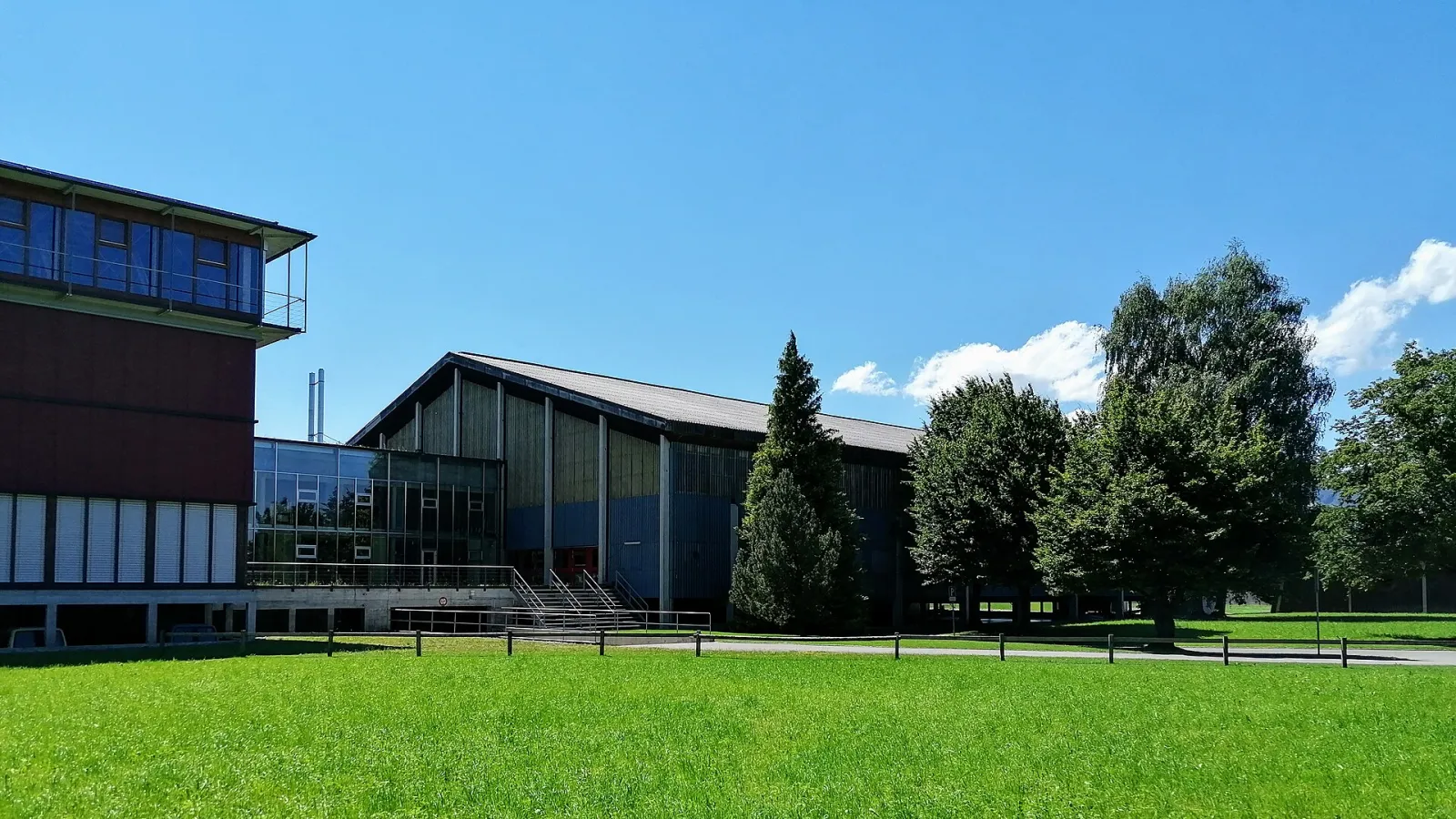 Die Eishalle im Aegeten in Widnau vom Park aus gesehen, mit dem Satteldach der Halle und dem angrenzenden Gebäude – Eishalle Rheintal