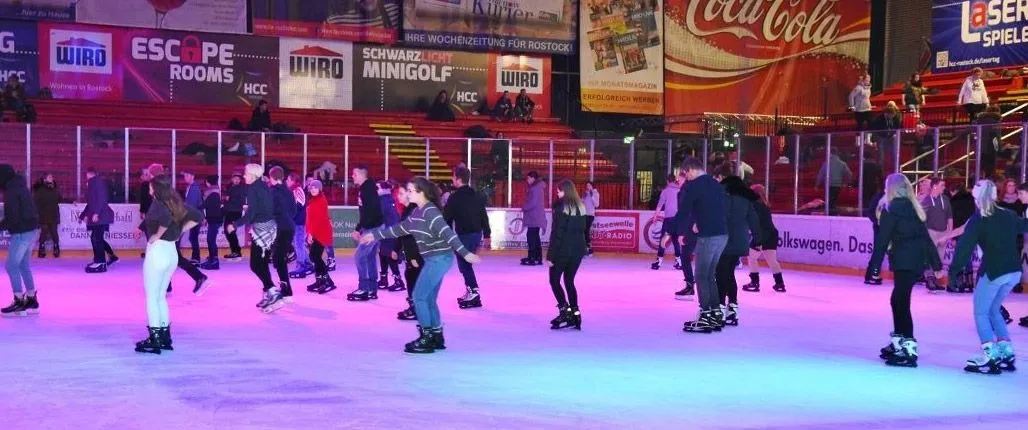 A public skating session in the Rostock ice hall, skaters scattered across the pink-lit ice with advertising boards along the far side. — Eishalle Rostock