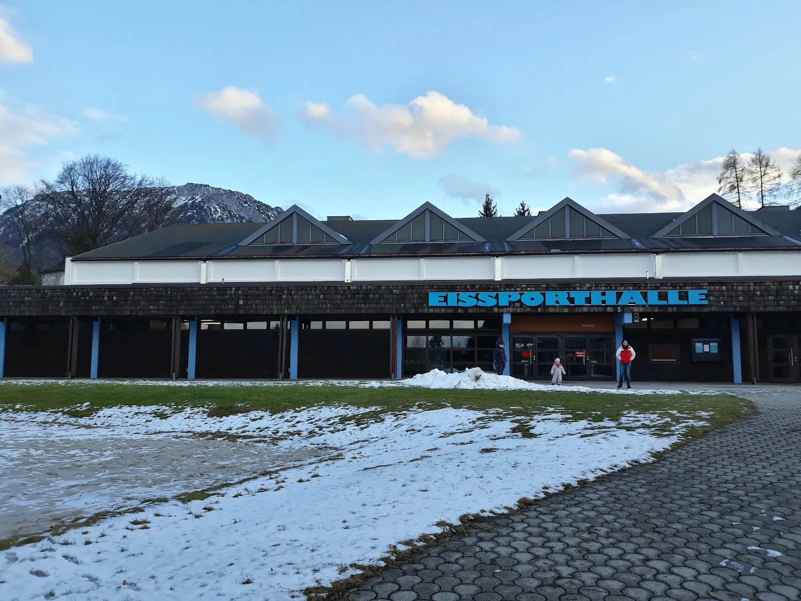 Die lange, dunkel verkleidete Front der Halle mit dem blauen Schriftzug EISSPORTHALLE und einer Reihe von Giebeln, dahinter Berge und davor ein Schneefleck auf der Wiese. – Eishalle Ruhpolding