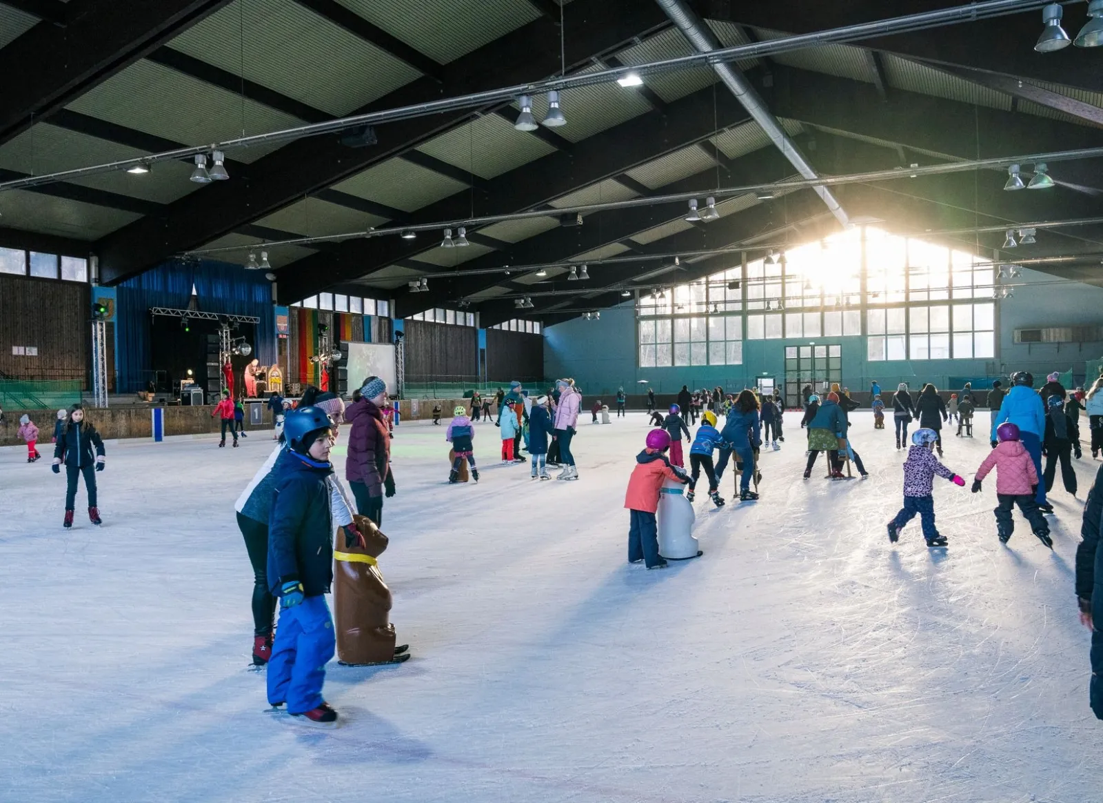 Schlittschuhläufer jeden Alters auf dem Eis der Halle Ruhpolding, Sonnenlicht fällt durch die hohen Fenster unter dem dunklen Stahldach. – Eishalle Ruhpolding