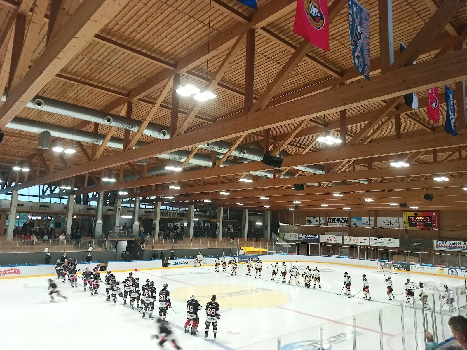 Weiter Blick über das Eis der Eishalle Sagibach in Wichtrach, mit einer Mannschaft an der gegenüberliegenden Bande, dem Holzdach und den Rängen darüber. – Eishalle Sagibach