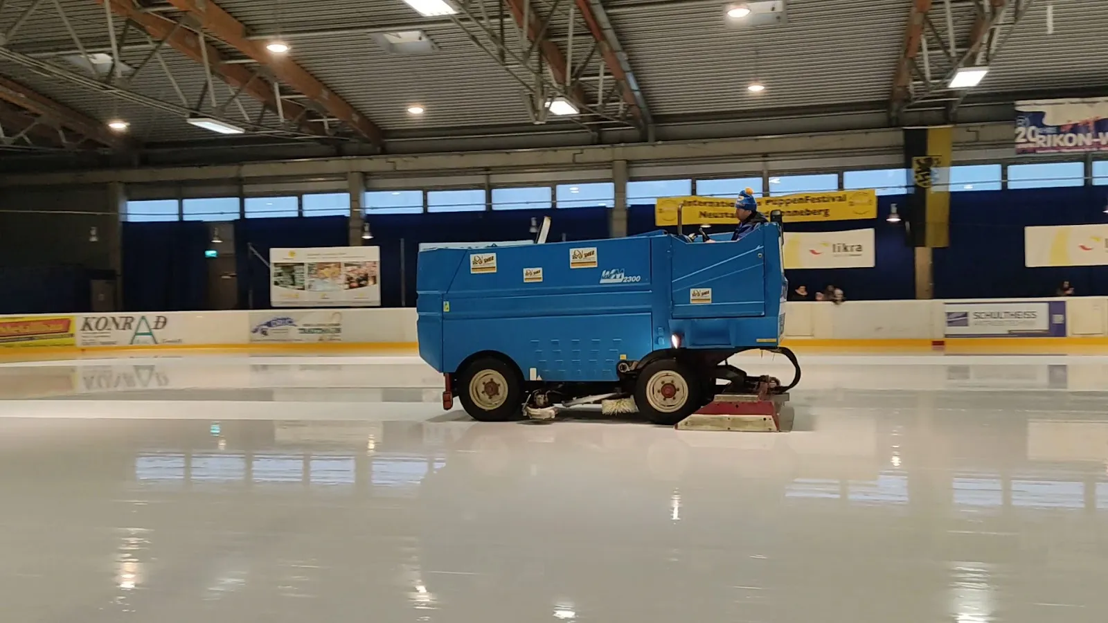 The clean ice sheet with a blue ice resurfacer parked mid-rink, sponsor boards along the far side and timber roof trusses above. — Eishalle Sonneberg (Eiswelt, SonneBad)