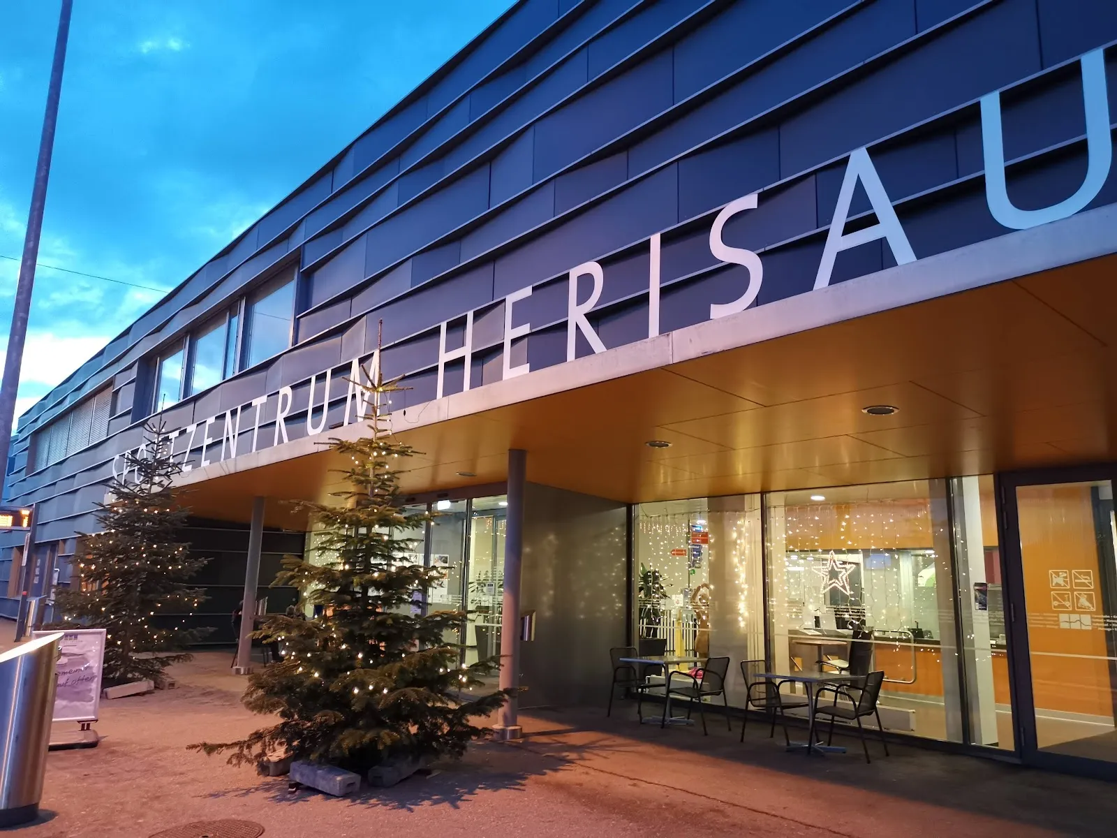 Der verglaste Eingang des Sportzentrums Herisau in der Abenddämmerung, mit dem Namen in grossen Buchstaben entlang der dunklen Metallfassade und beleuchteten Bäumen an der Tür – Eishalle Sportzentrum Herisau