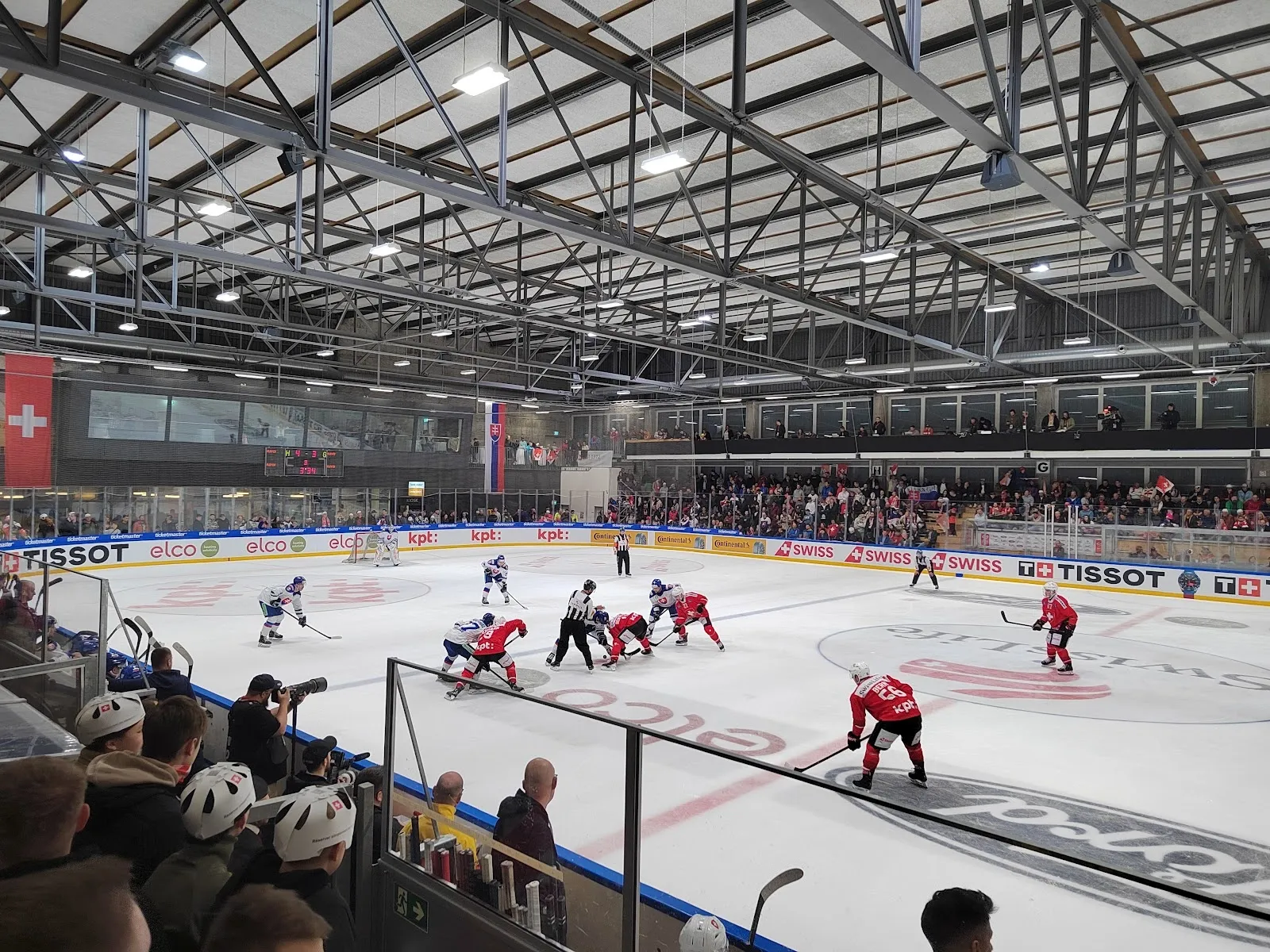 Ein Eishockeyspiel auf dem Eis des Sportzentrums Herisau, von den Rängen über die ganze Fläche gesehen, darüber der Stahlrahmen des Dachs. – Eishalle Sportzentrum Herisau