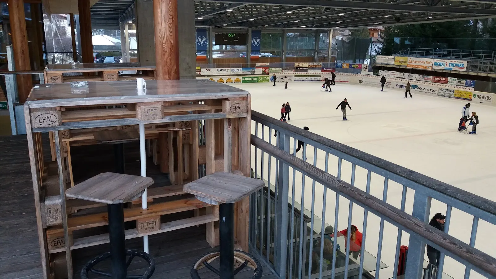 La pista coperta di Dobbiaco vista oltre la ringhiera di una terrazza in legno, con alcuni pattinatori lontani sul ghiaccio e i cartelloni degli sponsor lungo il lato opposto – Eishalle Toblach (Palaghiaccio Dobbiaco)