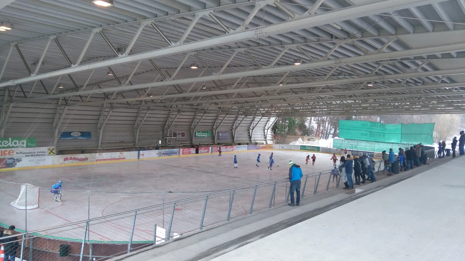 Weiter Blick über das Eis der Mehrzweckhalle Velden mit Schlittschuhläufern und einer Eishockeygruppe auf dem Eis, dem Stahldach und den Rängen hinter der Bande. – Eishalle Velden (Mehrzweckhalle)