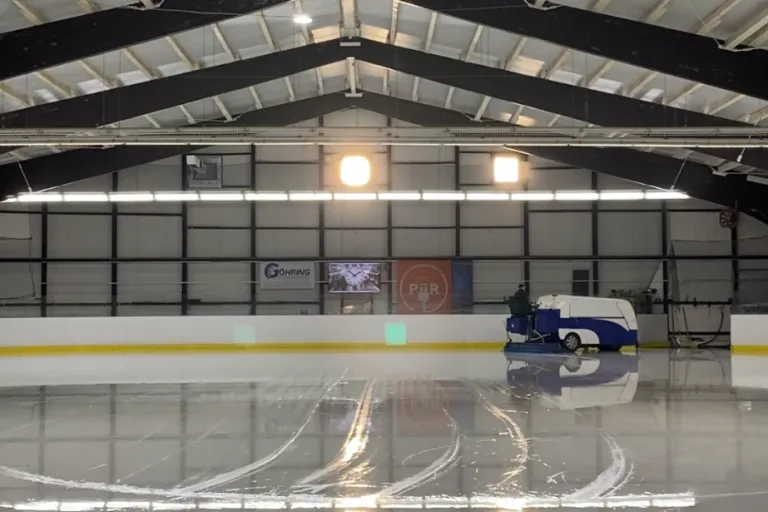 Inside the Eishalle Wabern: a smooth ice surface under the steel roof, with a resurfacing machine at the far end and boards with advertising along the side. — Wintersport-Center Wabern