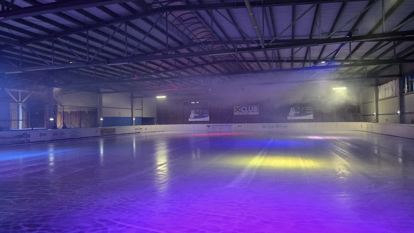 The indoor ice hall under a steel roof with the rink lit in purple and blue, the boards and sponsor panels along the far wall; no people on the ice. — Eishalle Waidhofen an der Ybbs