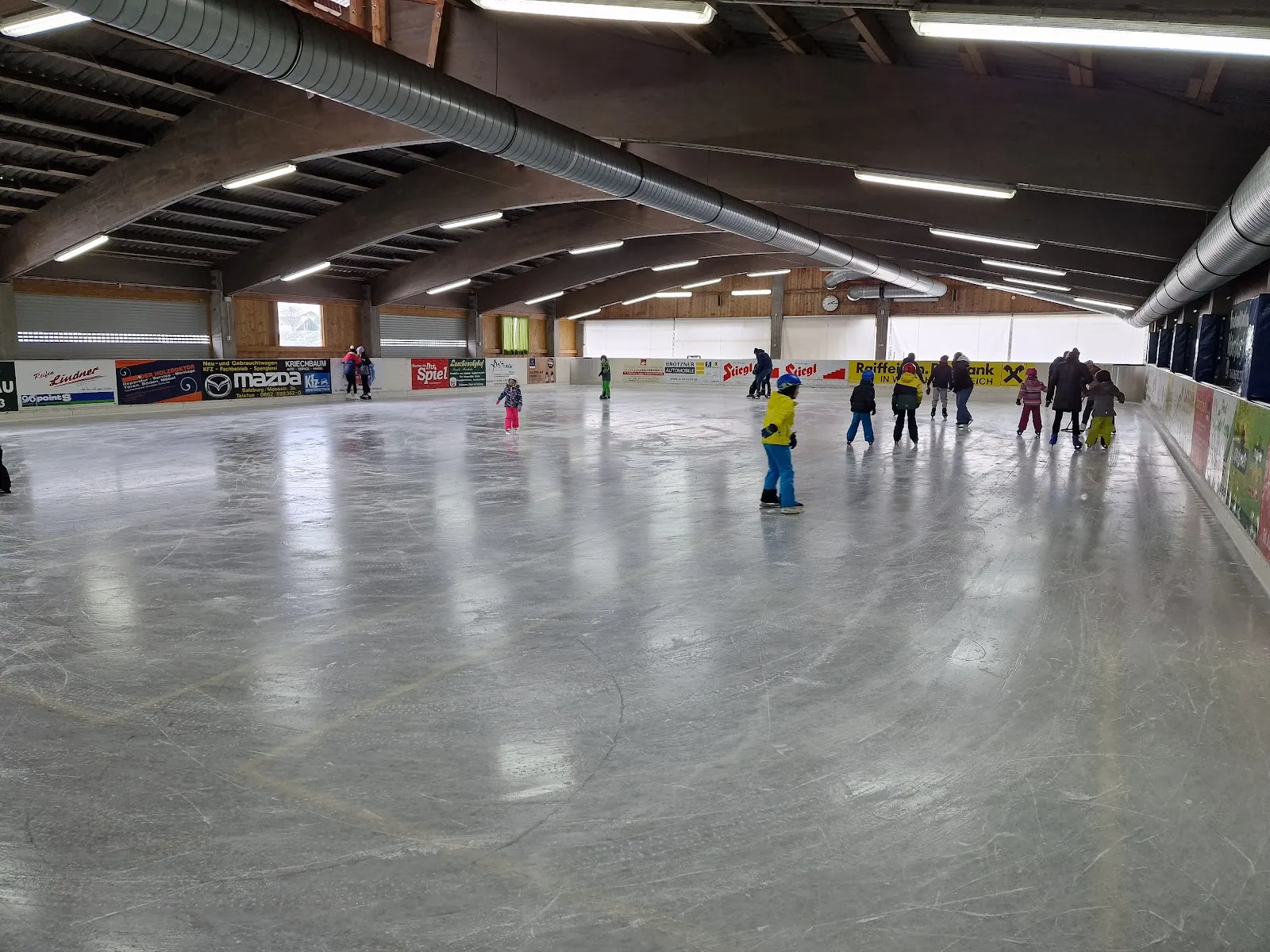 Die Eishalle mit ihrem Holzdach und der weiten grauen Eisfläche, ein paar kleine Gestalten weit entfernt am anderen Ende und die Banden an der Seite. – Eishalle Wals/Grünau
