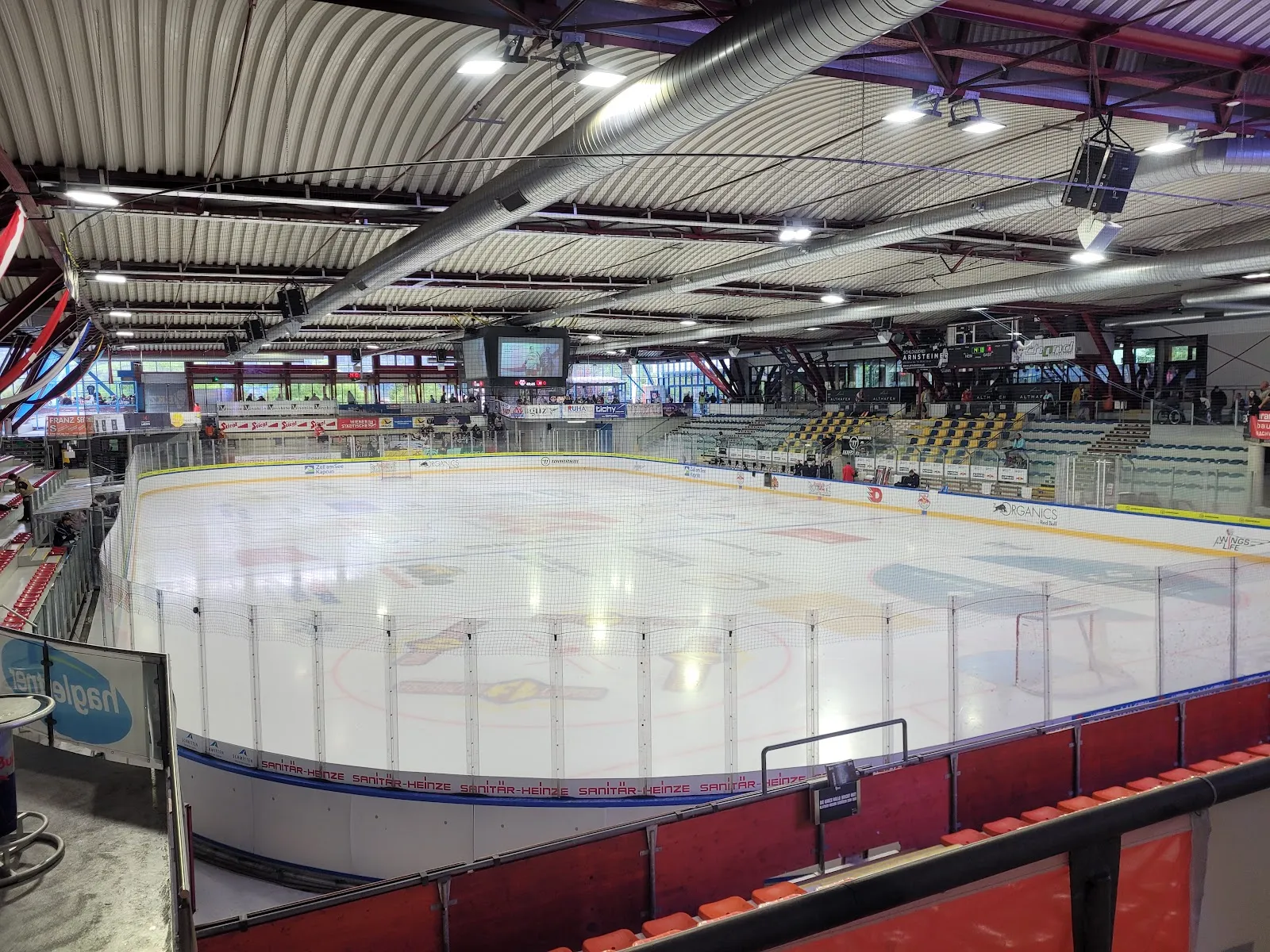 The empty ice sheet of the Eishalle Zell am See seen from the upper stand, under the curved steel roof with the red-edged boards and the seating around it — Eishalle Zell am See