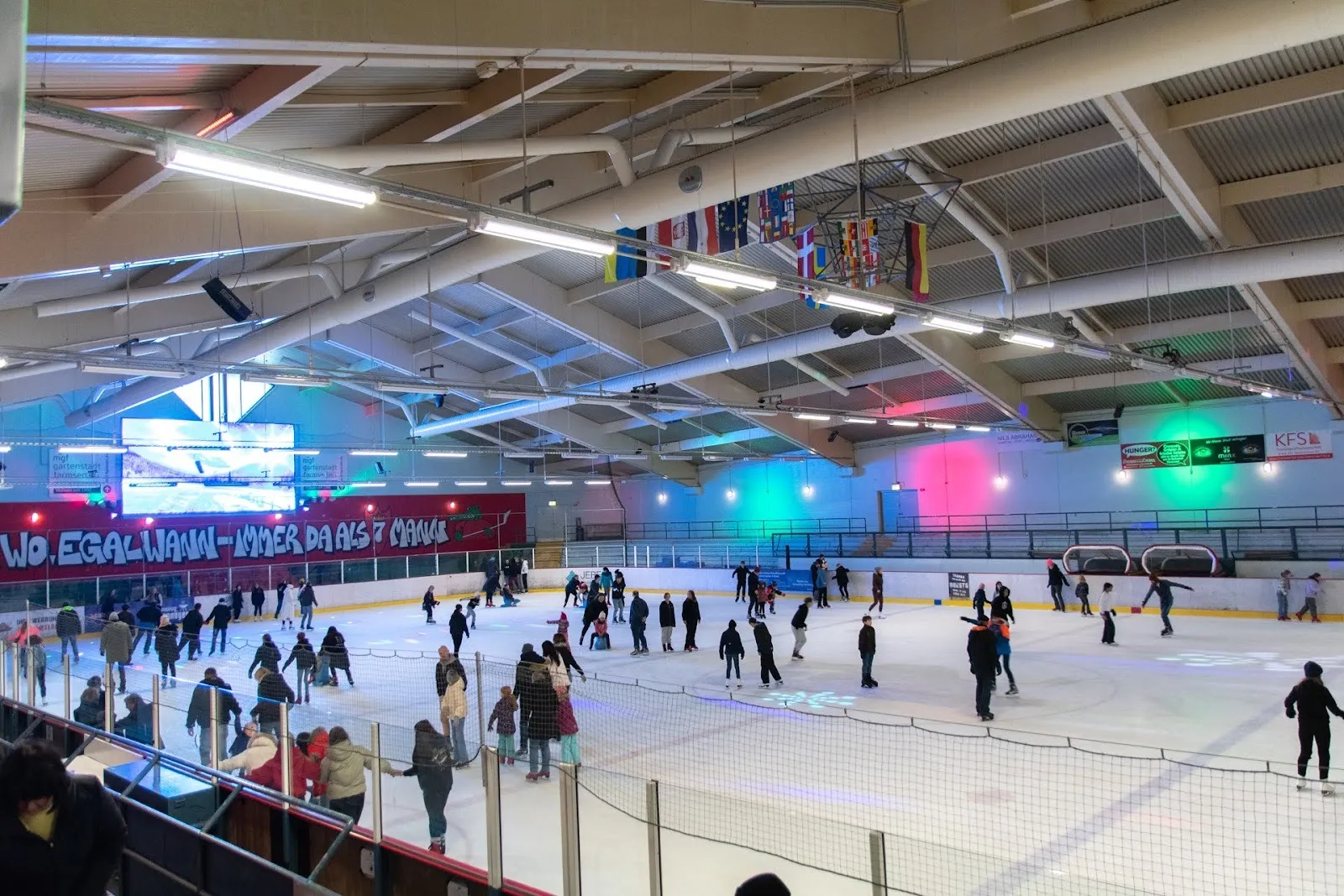A public skating session in the Eisland Farmsen hall, skaters spread across the ice under coloured lights and the steel-trussed roof. — Eisland Farmsen