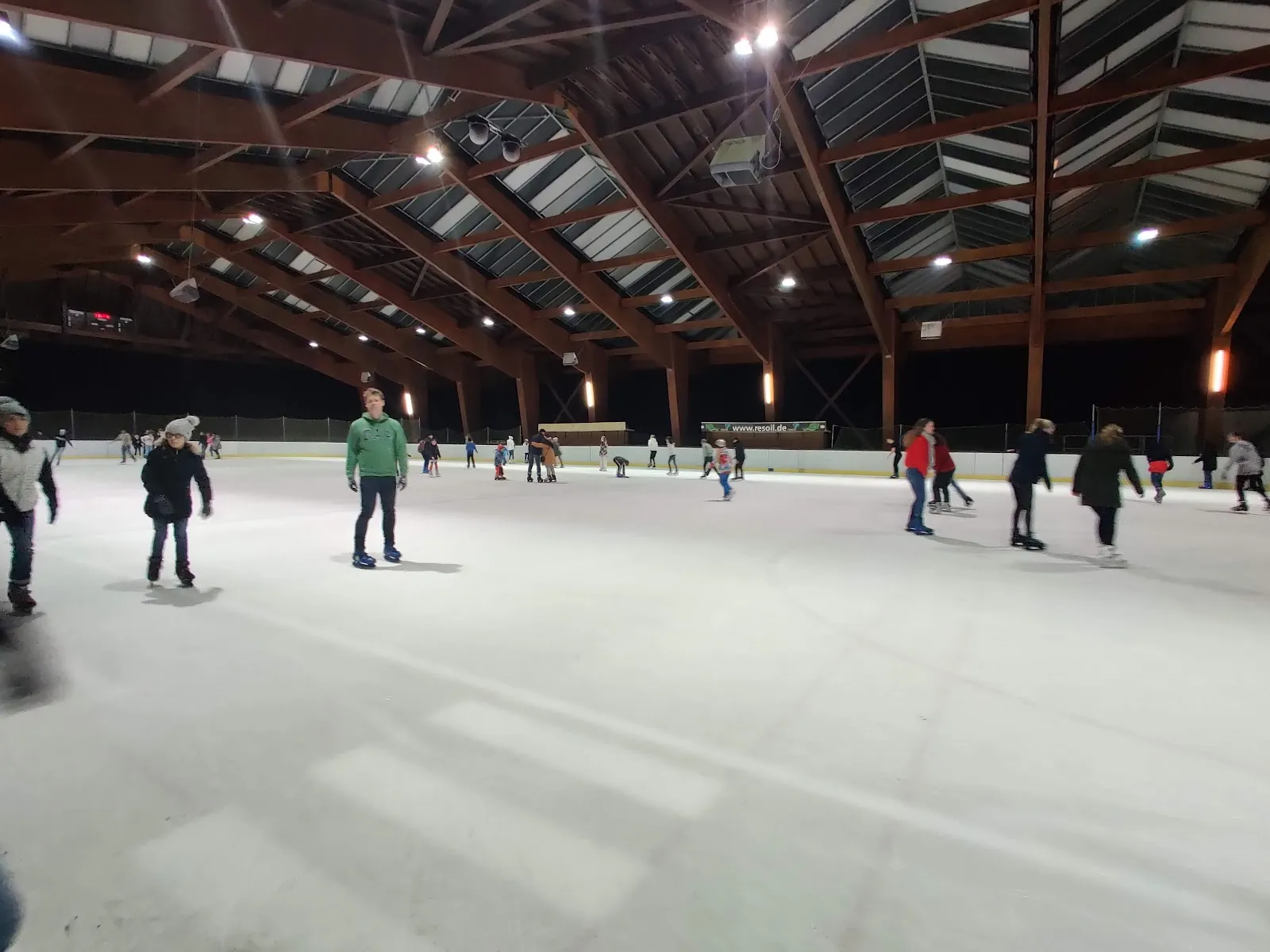 Public skaters spread across the ice under the roof's wooden trusses, with the lights on and the rink's edge dark beyond them. — Eislaufanlage Illerau