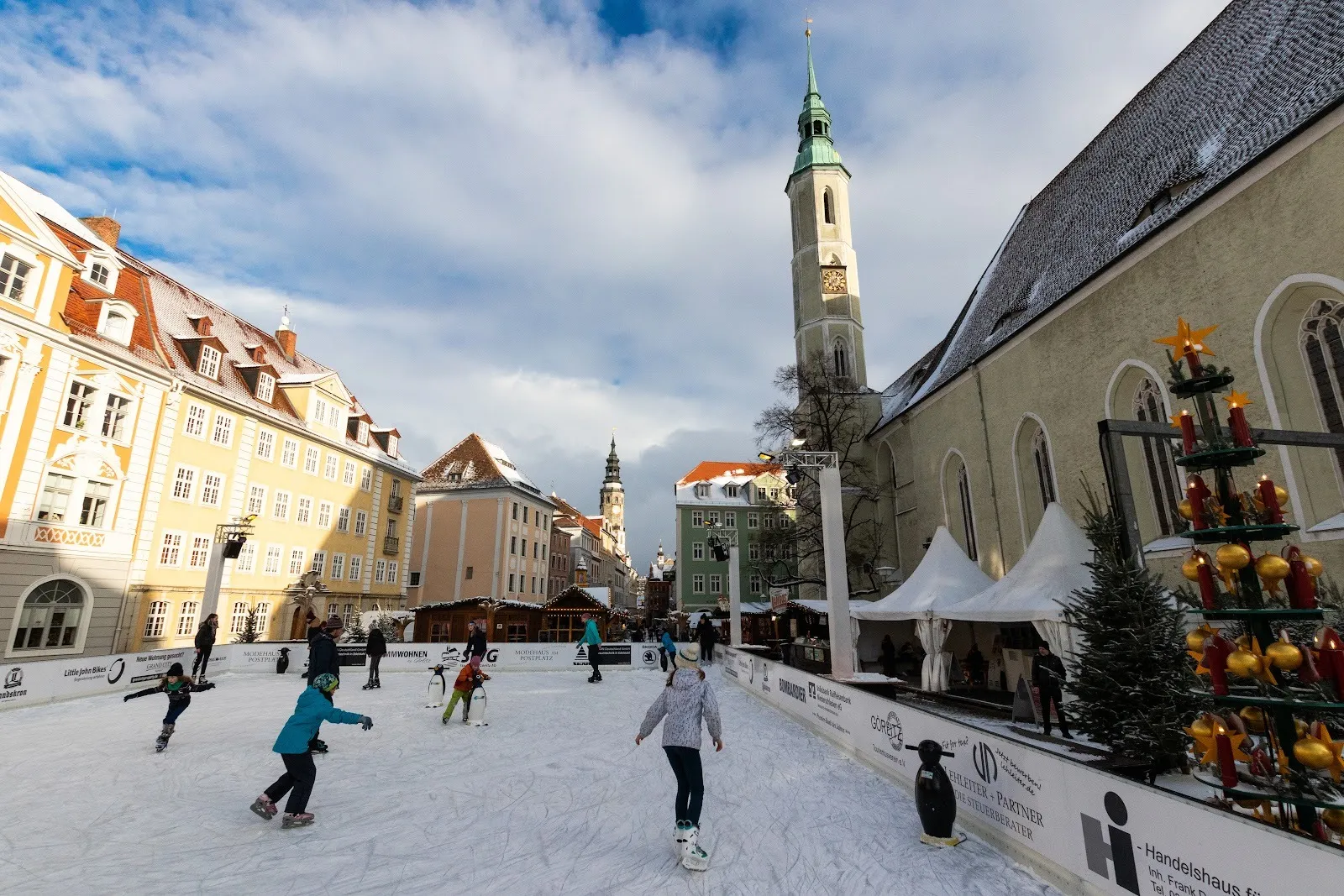 Schlittschuhläufer auf der temporären Eisbahn auf dem Görlitzer Altstadtplatz, dahinter Kirchturm und Marktstände. – Eislaufbahn Görlitz