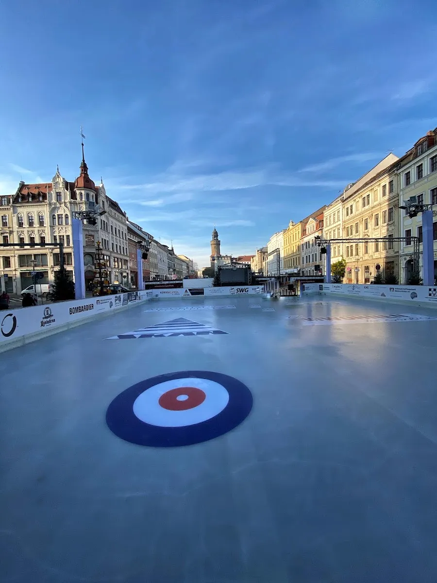 Die provisorische Eisbahn entlang des Obermarkts, leer, mit einer auf das Eis gemalten Curling-Zielscheibe und den historischen Bürgerhäusern und dem Turm dahinter unter blauem Himmel. – Eislaufbahn Görlitz
