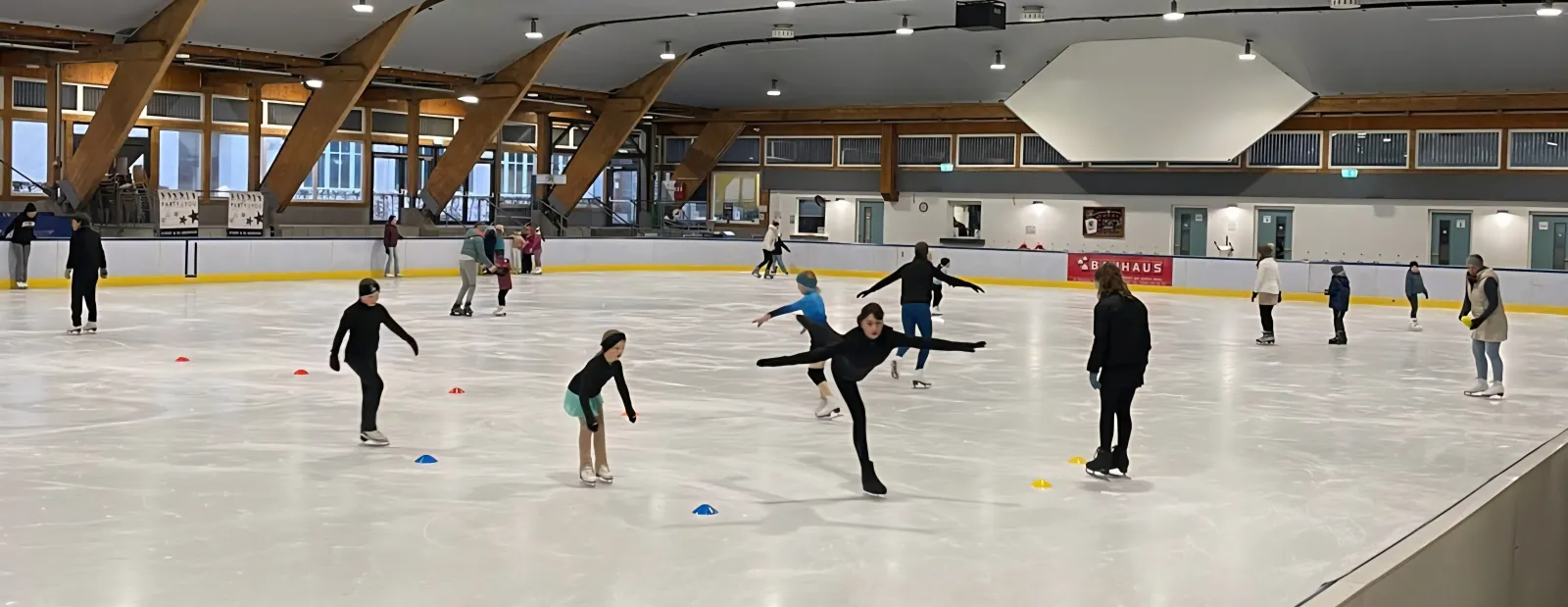 Skaters practising on the ice of the Baiersbronn hall under its timber-framed ceiling, with the long pale boards behind. — Eislaufhalle Baiersbronn