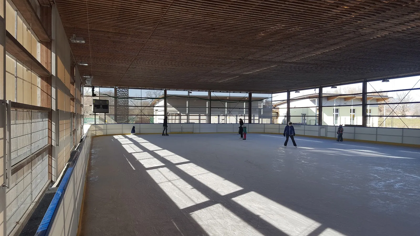 Die überdachte Eisbahn mit ihren langen geraden Banden und der Holzdecke; einige wenige Personen weit hinten am anderen Ende. – Eislaufhalle Bergxi