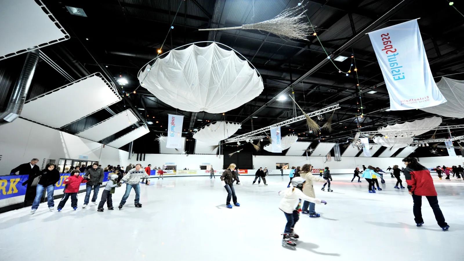 Die Eisfläche der Eislaufhalle Offenburg mit weißen Deckenelementen und Fahnen, Schlittschuhläufer in der Ferne – Eislaufhalle Offenburg
