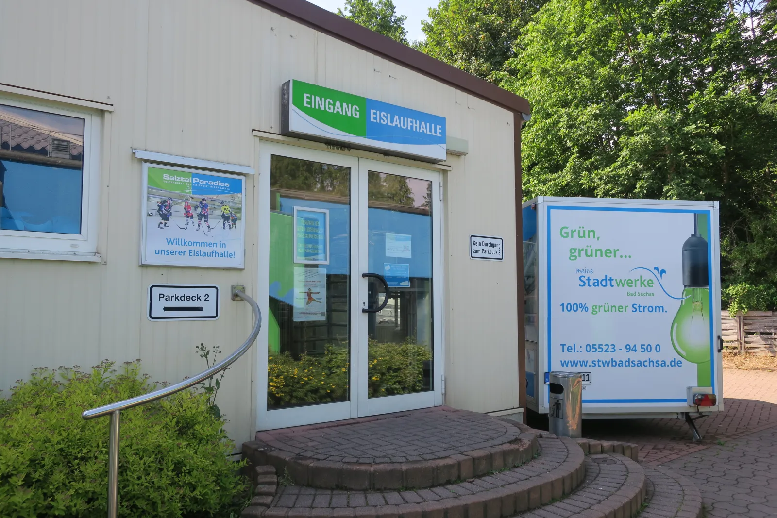 Der Eingang der Eislaufhalle in einem niedrigen cremefarbenen Gebäude mit grün-blauem Schild "Eingang Eislaufhalle", einem Willkommensplakat, Backsteinstufen und Bäumen dahinter. – Eislaufhalle Salztal Paradies