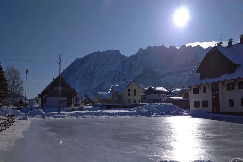 The smooth ice of the Bad Mitterndorf rink in low winter sun, with village houses and a mountain ridge behind. — Eislaufplatz Bad Mitterndorf