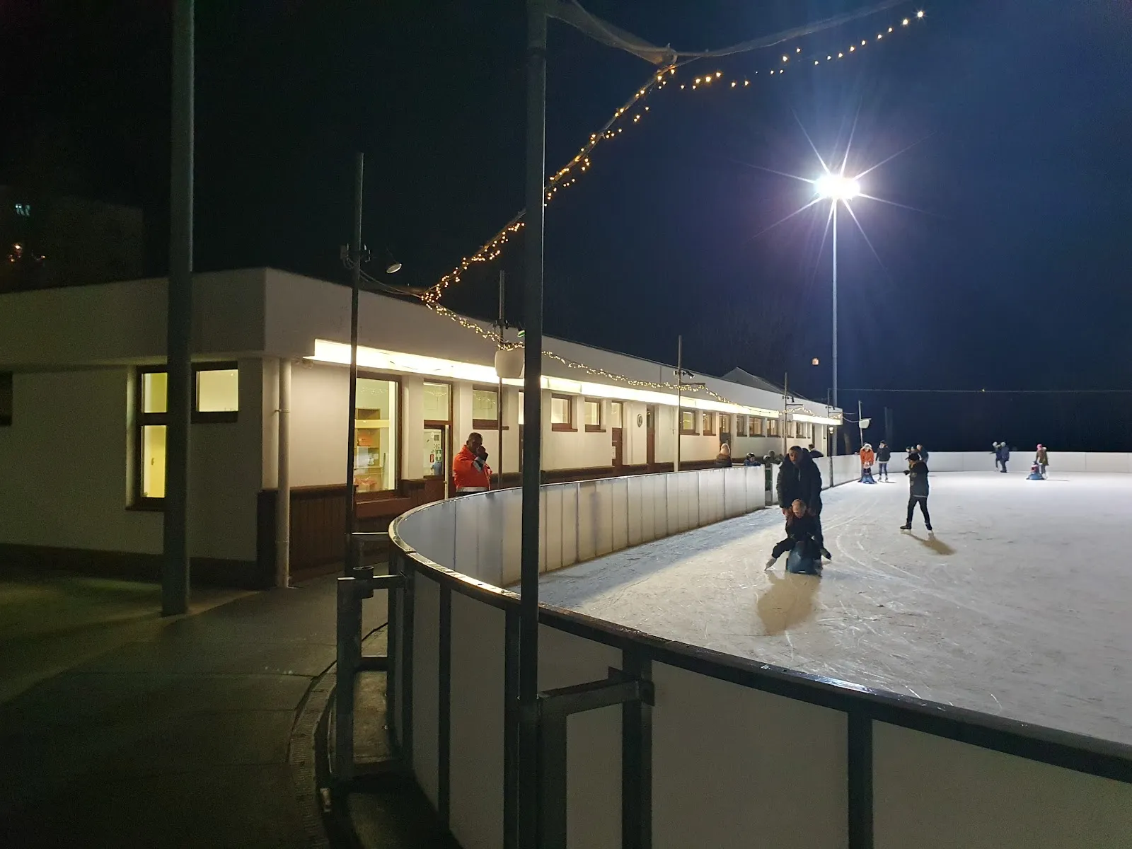Night view of the Schwechat rink from its edge, with the single-storey clubhouse with lit windows along the rink. — Eislaufplatz Freizeitzentrum Schwechat