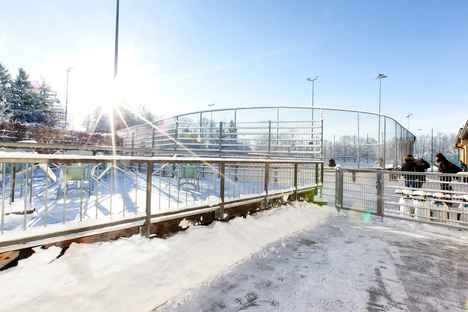 Der umzäunte Rand der Eisbahn und die verschneiten Zuschauerstufen im tiefen Winterlicht, hinter dem Netz ragen Flutlichtmasten auf. – Eislaufplatz Grünwalder Freizeitpark