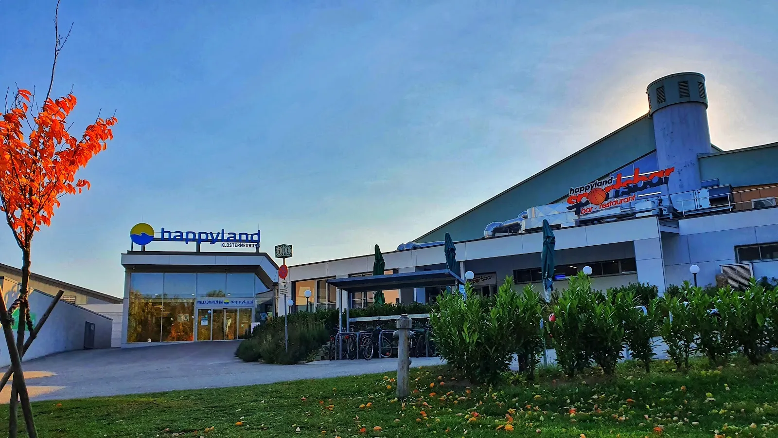 Die Front des Happyland-Gebäudes am Abend, mit dem Happyland-Schriftzug an der Fassade sowie den Hecken und dem Eingang davor. – Eislaufplatz Happyland Klosterneuburg