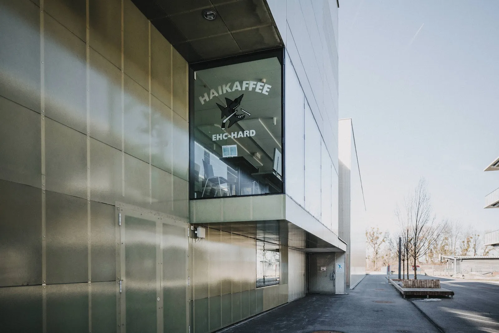 Die Front des Eisbahngebäudes am Seeweg: graues Metall und Glas mit den Schildern von Haikaffee und EHC Hard im Fenster. – Eislaufplatz Hard