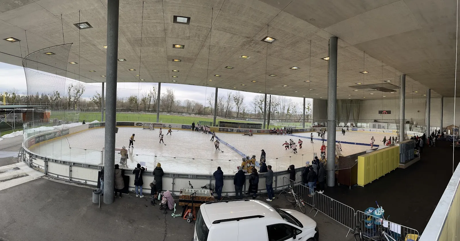 Die überdachte Freiluft-Eisbahn in der Weitansicht, mit dem Betondach darüber und der Eisfläche quer durch die Halle, Schlittschuhläufer klein in der Ferne. – Eislaufplatz Hard