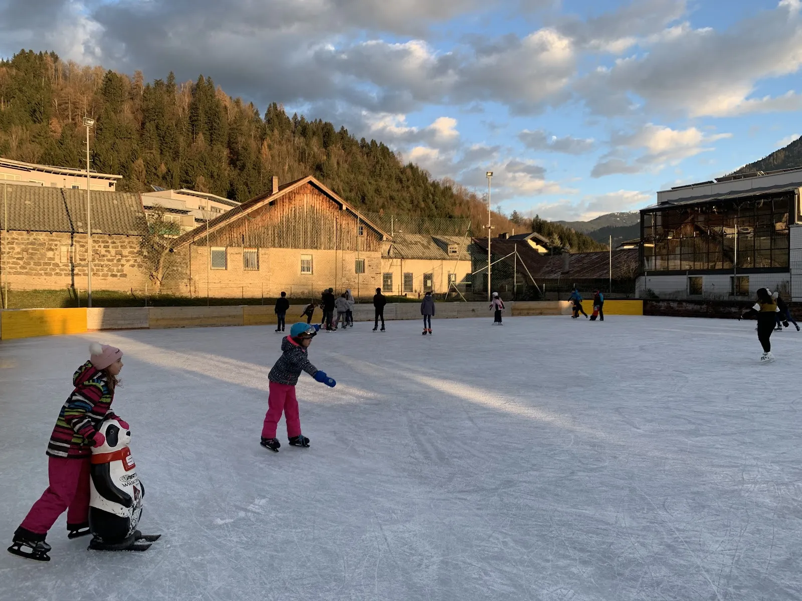 Kinder laufen im Licht des späten Nachmittags auf dem Freiluft-Eis; dahinter die niedrige Bande, Holzgebäude und ein bewaldeter Hang. – Eislaufplatz Jenbach