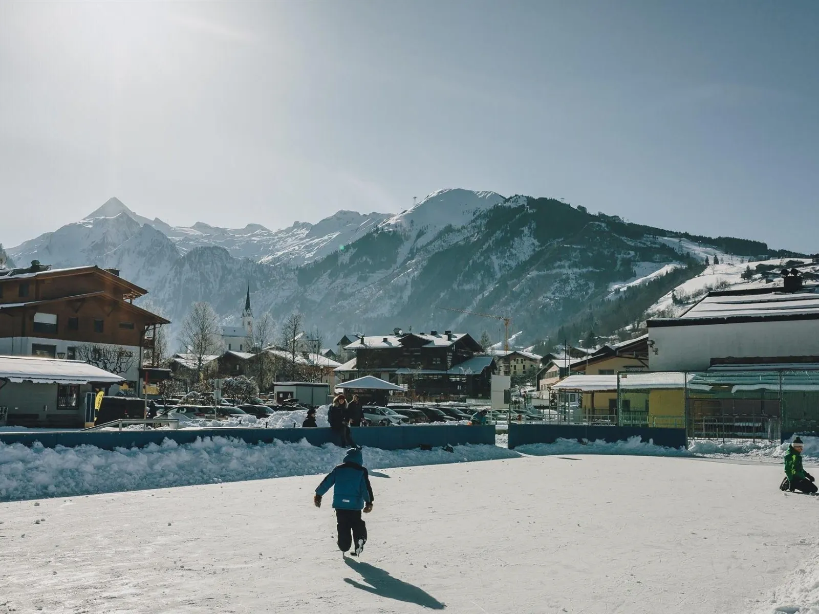 A child skating across the Kaprun rink, with the village rooftops and snow-capped mountains behind the boards. — Eislaufplatz Kaprun