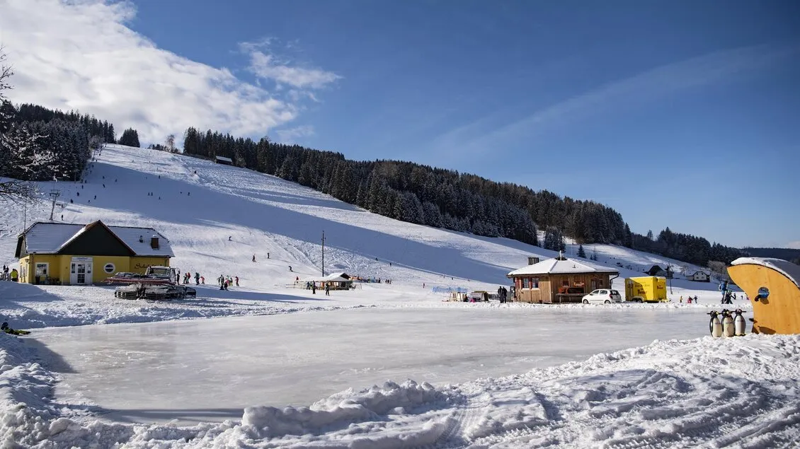 The Kleinlobming rink at the foot of the ski slope, with the lift hut and a yellow shelter beside the ice. — Eislaufplatz Kleinlobming