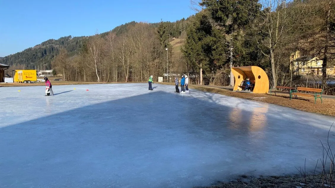 The frozen rink at Kleinlobming in winter sun, with skaters at the far side, a yellow wooden shelter and wooded hills behind. — Eislaufplatz Kleinlobming