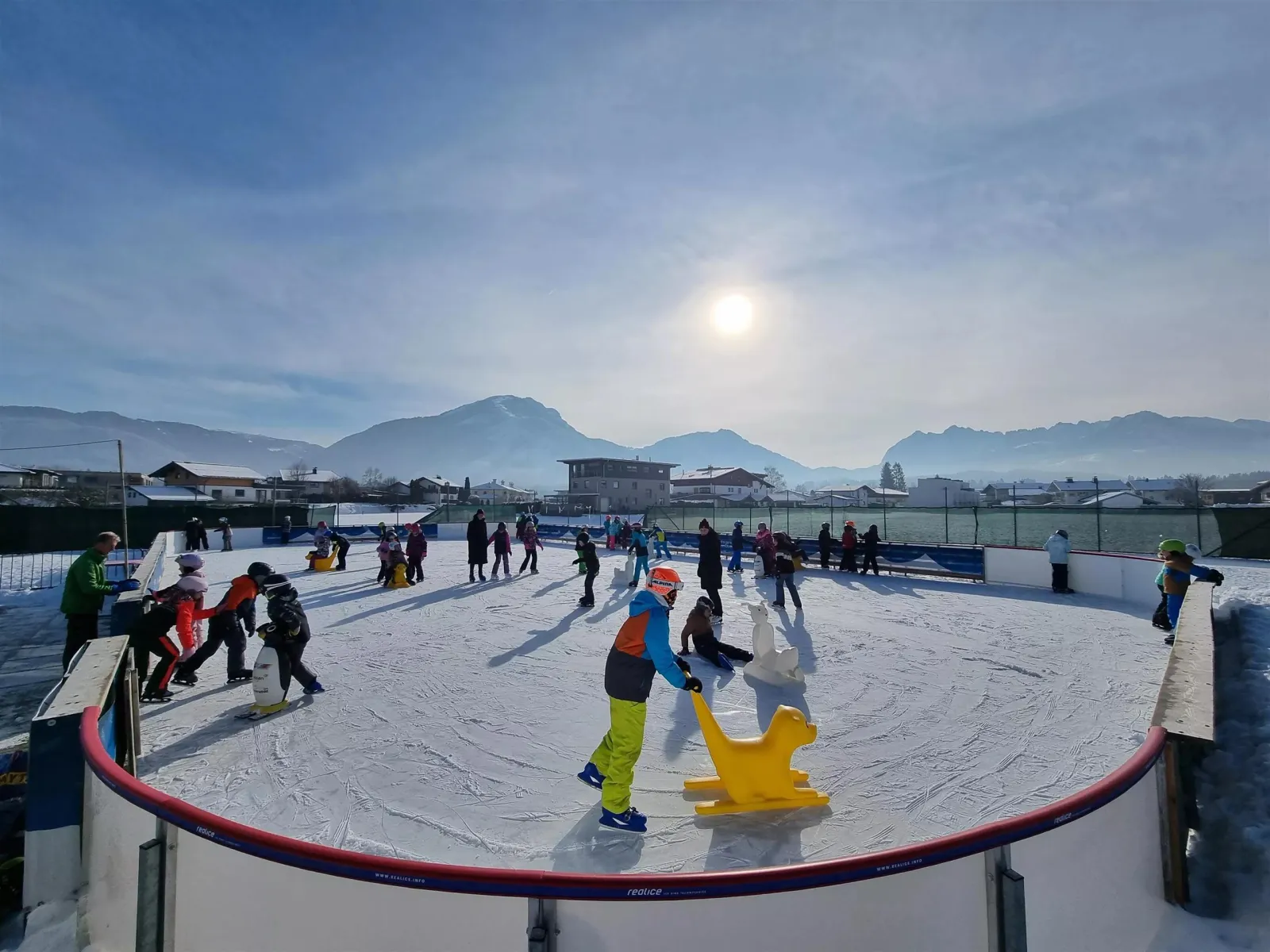 The outdoor artificial ice rink in Kössen on a sunny winter day, with children skating around the boards and the Kaiser mountains behind. — Eislaufplatz Kössen