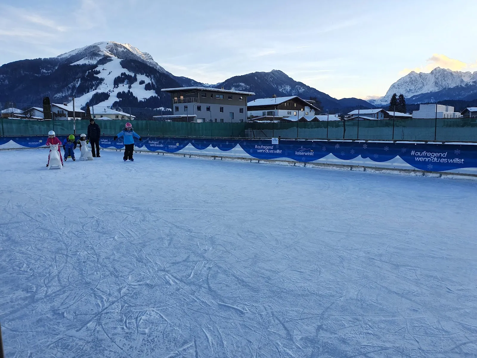 The Kössen ice-skating rink with a few skaters, mountains behind — Eislaufplatz Kössen